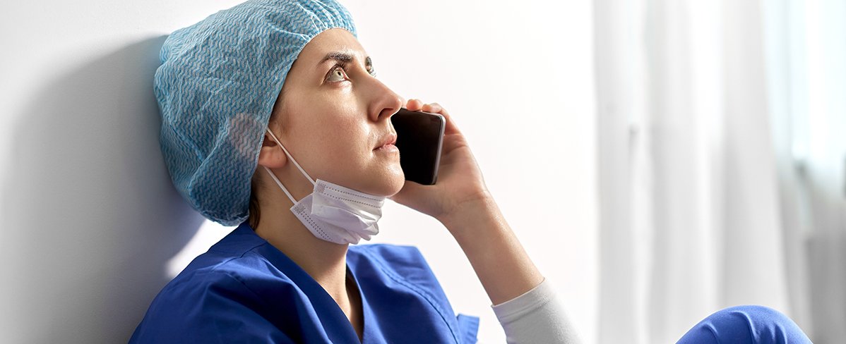 Traurige und müde junge Ärztin oder Krankenschwester, die auf dem Boden sitzt und ein Handy ans Ohr hält